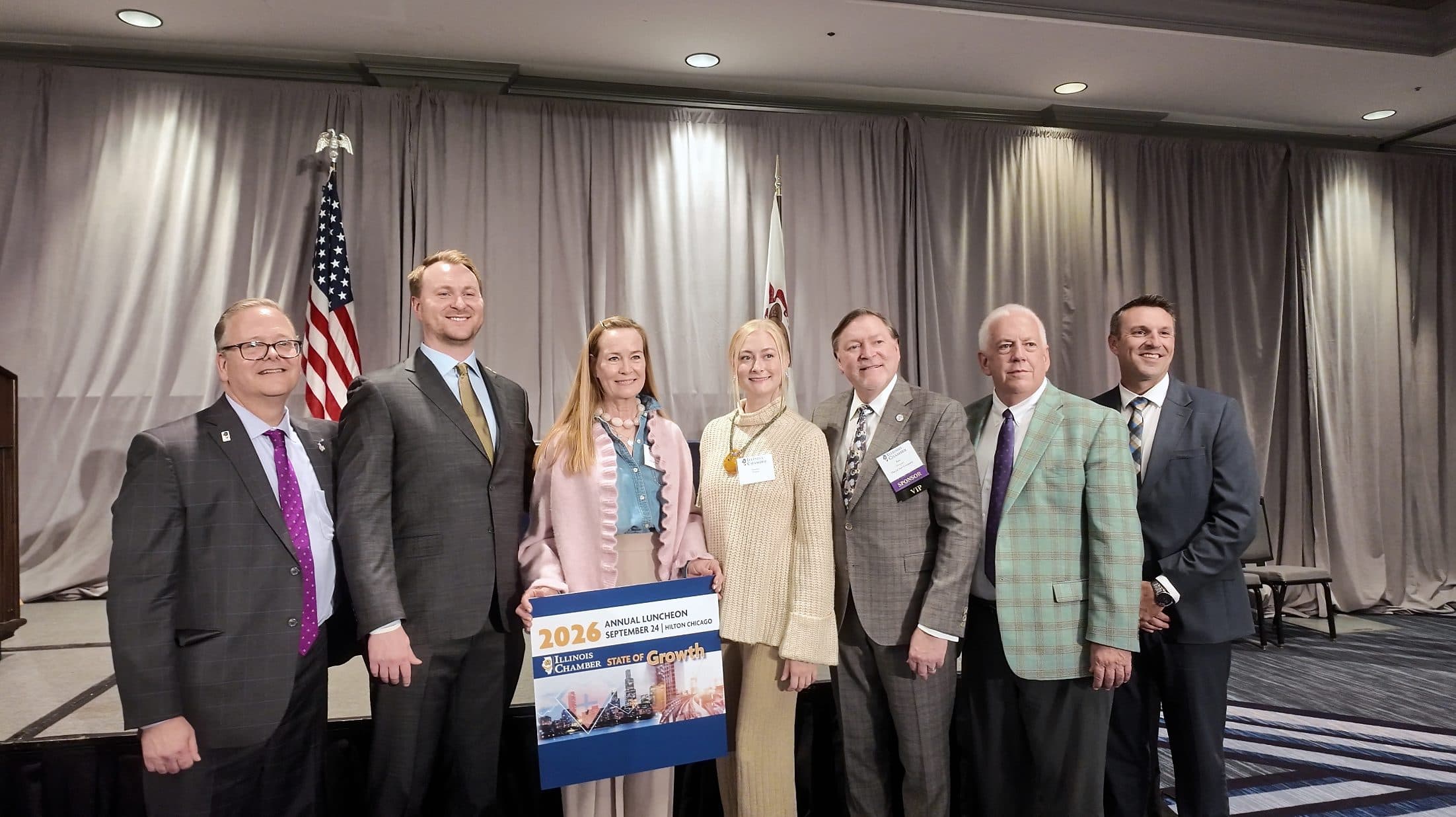 Seven people pose on stage with a 2026 Annual Luncheon sign.