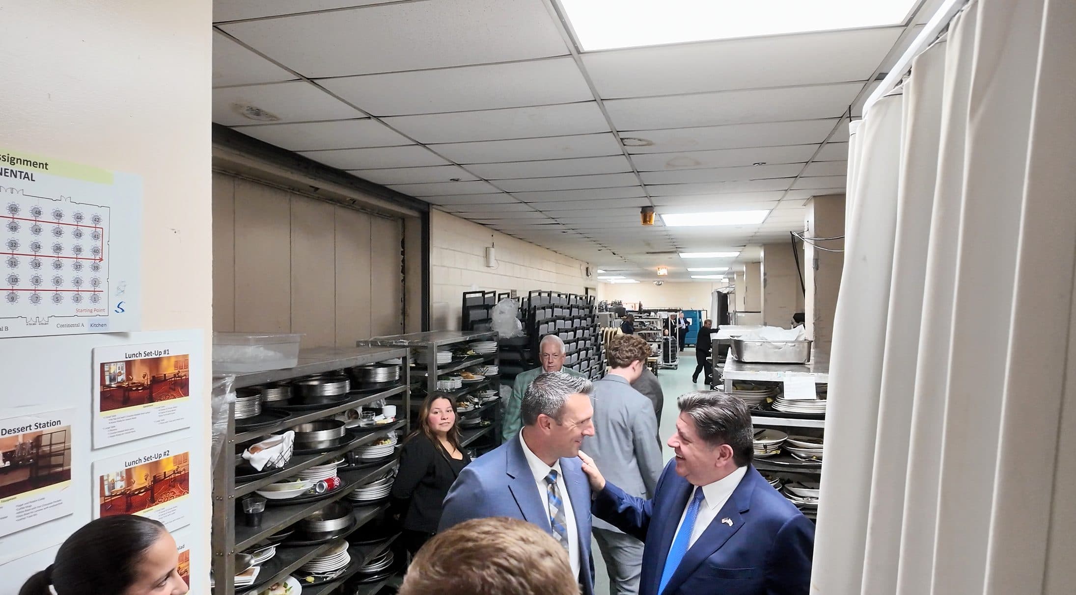 Two men talk in the backstage corridor, surrounded by guests and staff.