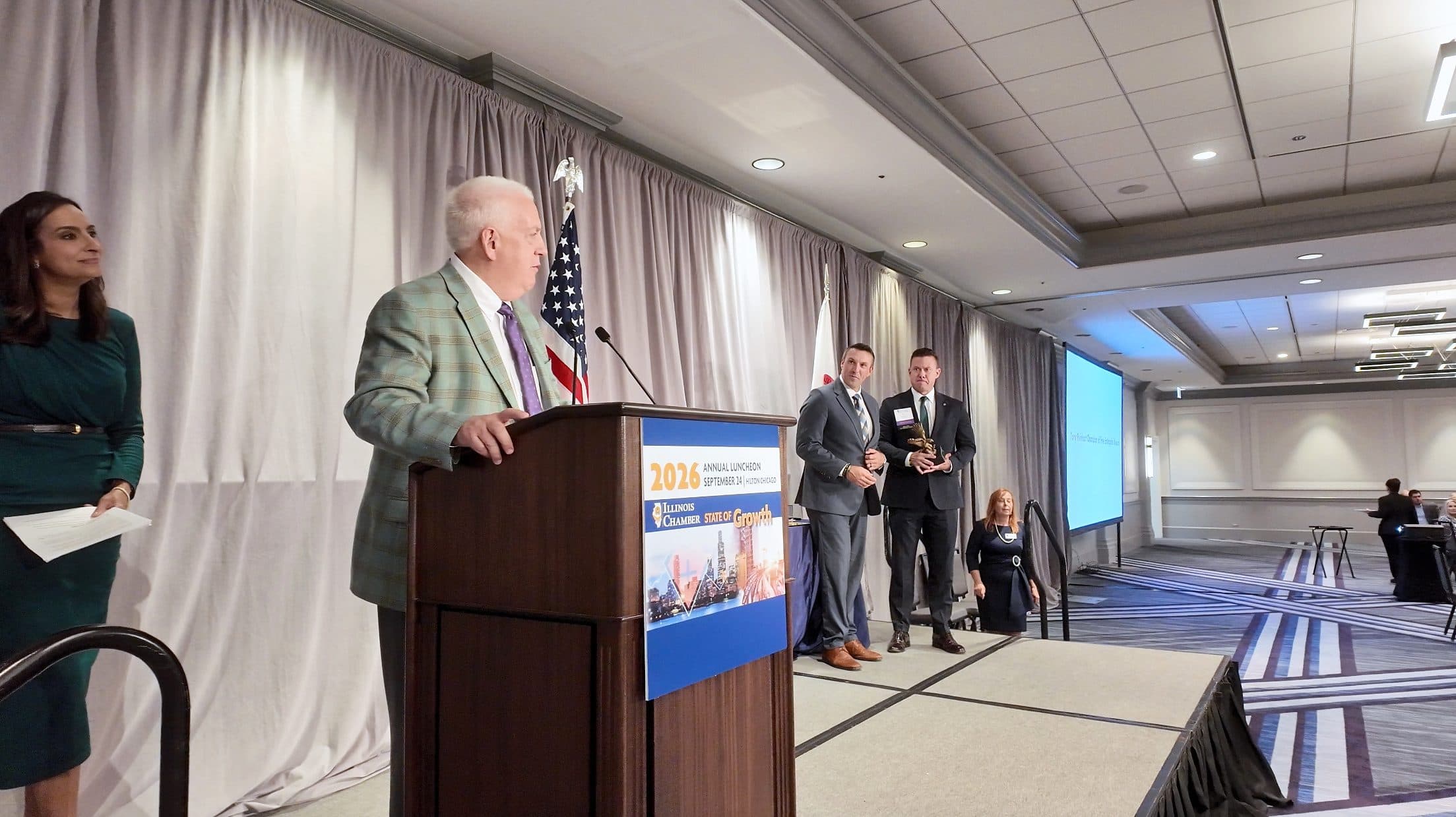 Award recipients hold their awards on stage as a speaker reads from the podium.